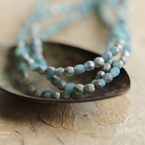 May include: A close-up of a multi-strand necklace made of light blue glass beads with silver accents. The necklace is resting on a rustic, brown surface.
