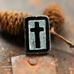 May include: A rectangular, black and teal pendant featuring a cross design. The pendant has a textured surface and is set against a rustic background. The text ".. arte bella.. 11239" is visible.