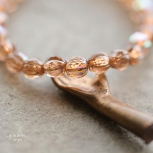 Può includere: Una collana delicata realizzata con piccole perle di vetro sfaccettate color pesca. La collana è infilata su un filo sottile e ha un design semplice ed elegante. La collana è appoggiata su una superficie testurizzata.