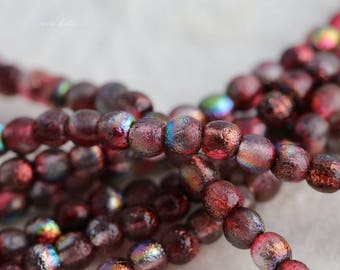 May include: Close-up of strands of iridescent, burgundy-colored beads. The beads have a textured surface and reflect multiple colors, including blue, green, and gold. The beads are round and appear to be made of glass or a similar material, suitable for jewelry making.