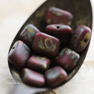 May include: Close-up of a collection of rectangular beads in a metal spoon. The beads are a deep burgundy color with patches of olive green and yellow. Each bead has a hole for stringing. The beads have a matte finish.