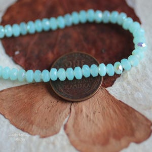May include: A light blue beaded bracelet made of faceted glass beads. The bracelet is lying on a brown and white surface.