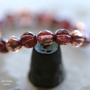 Peut inclure: Un bracelet en gros plan composé de perles de verre facettées et irisées dans des tons de rose et de cuivre. Les perles sont disposées en cercle et reposent sur un dé à coudre brun foncé.