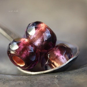 May include: Three faceted, dark purple beads rest in a silver spoon. The beads have a glossy finish and a mix of dark purple and reddish-brown hues. The beads are likely for jewelry making or crafting.