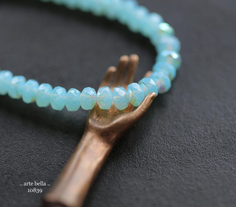 May include: A delicate bracelet made of light blue faceted glass beads. The bracelet is held by a hand-shaped bronze charm.