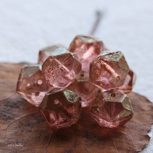 Op de afbeelding: Een verzameling gefacetteerde, roze glazen kralen. De kralen hebben een geometrische vorm met meerdere zijden en een gat om te rijgen. Ze liggen op een bruin blad.