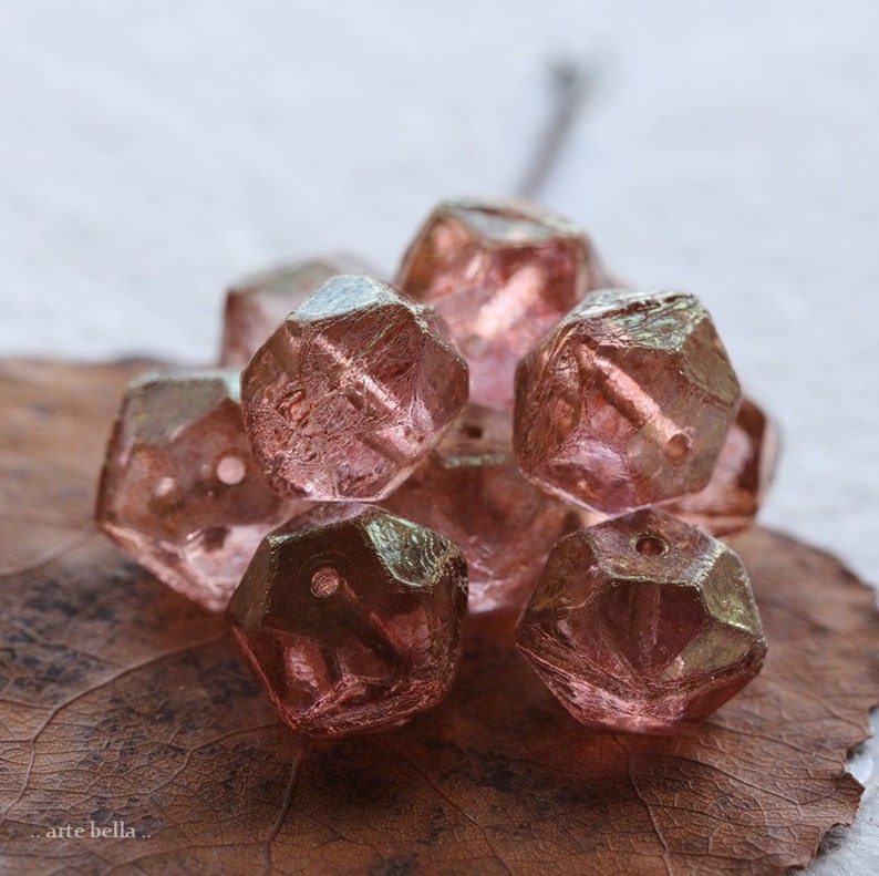 Op de afbeelding: Een verzameling gefacetteerde, roze glazen kralen. De kralen hebben een geometrische vorm met meerdere zijden en een gat om te rijgen. Ze liggen op een bruin blad.