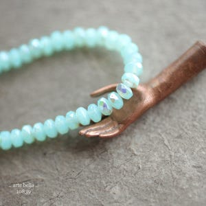 May include: A light blue crystal bead necklace with a copper hand charm. The necklace is made of faceted beads and the hand charm is detailed with a textured finish.