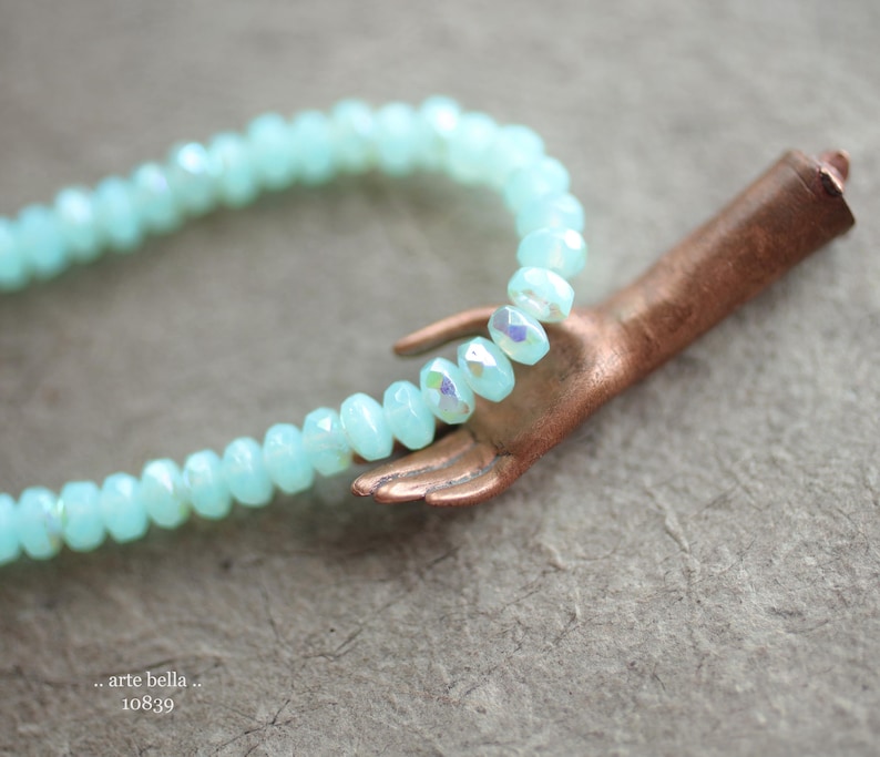 May include: A light blue crystal bead necklace with a copper hand charm. The necklace is made of faceted beads and the hand charm is detailed with a textured finish.