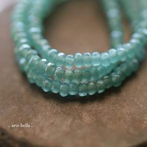 May include: A close-up of a strand of light blue gemstone beads. The beads are faceted and have a slightly irregular shape. The beads are strung on a clear thread and are arranged in a circular pattern.