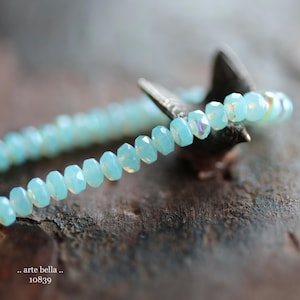 May include: A delicate bracelet made of light blue faceted beads. The bracelet is strung on a silver wire and is resting on a piece of wood.