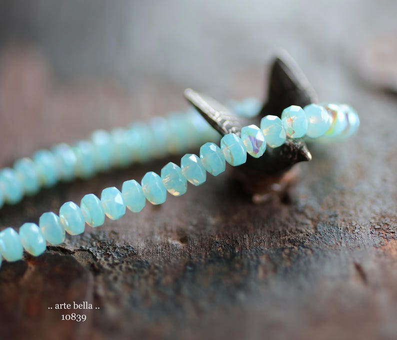 May include: A delicate bracelet made of light blue faceted beads. The bracelet is strung on a silver wire and is resting on a piece of wood.