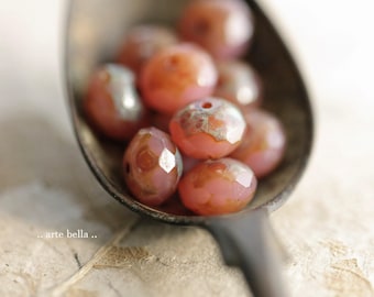 May include: A collection of round, faceted pink glass beads with metallic silver-toned accents resting in a tarnished metal spoon. The beads feature a translucent finish and a central hole for stringing. The text .. arte bella .. is visible.