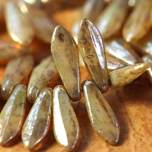 May include: Close-up of several elongated, iridescent beads in shades of gold and amber. The beads have a smooth, glossy surface and a teardrop shape. The beads are scattered, creating a textured, shimmering effect. The beads are likely for jewelry making.