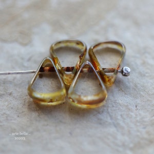 May include: Close-up of four amber-colored glass teardrop-shaped pendants, connected by a thin, rusty-colored metal bar. The pendants are suspended on a silver-colored wire. The text ".. arte bella .." and "10203" are visible.