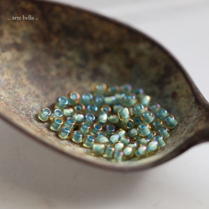 May include: A close-up of a pile of small, iridescent teal and gold glass beads in a vintage metal scoop. The beads have a slightly irregular shape and a subtle shimmer.