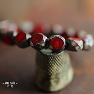 May include: A close-up of a red glass bead bracelet with a metallic sheen. The bracelet is resting on a vintage-looking thimble.