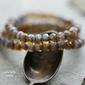 May include: Two beaded bracelets with faceted, iridescent beads in shades of gold, brown, and lavender. The bracelets are resting on a silver spoon. The text ". arte bella ." and "10085" are visible.