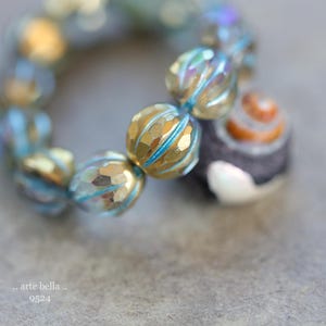 May include: A close-up of a bracelet with faceted gold and blue beads. The bracelet has a mix of colors, including gold, blue, and iridescent hues. The bracelet also features a dark gray bead with a spiral design and a white accent. The text ".. arte bella .. 9524" is visible.