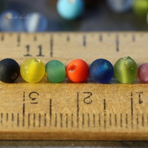 May include: A row of eight colorful glass beads, including black, yellow, green, orange, blue, and purple, lined up on a wooden ruler.