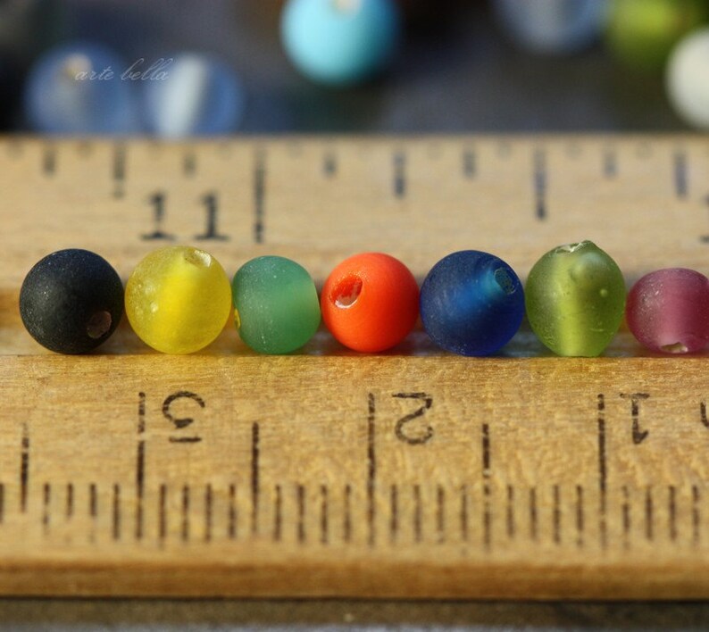 May include: A row of eight colorful glass beads, including black, yellow, green, orange, blue, and purple, lined up on a wooden ruler.