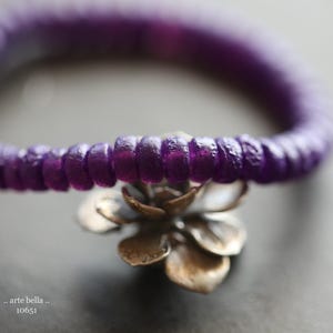 May include: A bracelet composed of small, circular, deep purple beads with a textured surface. A metallic, silver-toned flower charm with layered petals is attached to the centre of the bead strand. The text ".. arte bella .. 10651" is visible.