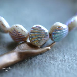 May include: A strand of iridescent white shell beads with a subtle pink and blue shimmer. The beads are arranged in a circular pattern and have a slightly textured surface.