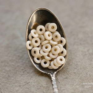 以下が含まれることがあります： 小さなオフホワイトのドーナツ型ビーズが山盛りになった銀のスプーン。ビーズは集まってスプーンを満たしています。スプーンには装飾的な柄があり、質感のある灰色の表面に置かれています。画像には「arte bella」と「11307」の文字が含まれています。