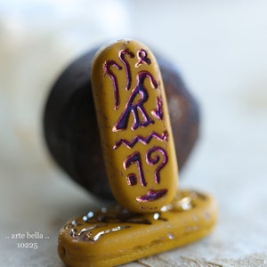 May include: A gold-colored glass bead with a purple design of ancient Egyptian hieroglyphs. The bead is sitting on a smaller, similar bead.