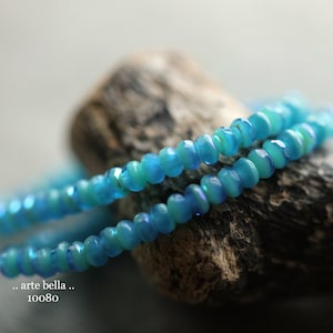 May include: Two strands of iridescent blue glass beads on a piece of driftwood. The beads have a light blue and green color.