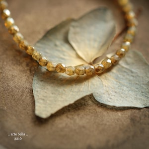 May include: A gold-toned beaded necklace with a delicate, faceted design. The necklace is draped over a dried flower, creating a natural and elegant aesthetic.