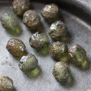 May include: A collection of green glass beads with a metallic finish. The beads are irregular in shape and have a textured surface.