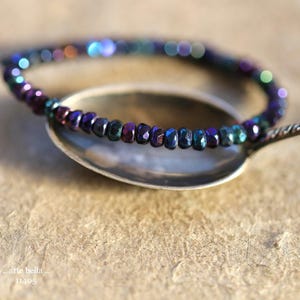 Könnte beinhalten: Ein kreisförmiges Perlenarmband mit kleinen, facettierten, irisierenden Perlen in dunklen Violett-, Blau- und Grüntönen. Das Armband liegt auf der Laffe eines silberfarbenen Metalllöffels. Der Text "arte bella .. 11495" ist in der Ecke sichtbar.