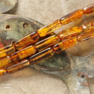 May include: Close-up of amber-colored rectangular beads with dark inclusions. The beads are strung together, showcasing their translucent quality. The background features a weathered, metallic surface. The text ".. arte bella .. 11464" is visible.