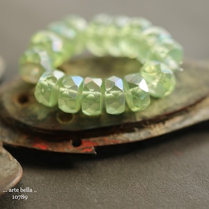 May include: A strand of light green glass beads with a faceted, geometric design. The beads are arranged in a circle and are resting on a textured, brown surface.
