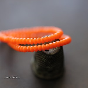May include: A close-up of a bright orange glass bead necklace wrapped around a small, dark gray thimble. The necklace is made of small, round beads.