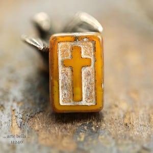 May include: An amber-coloured rectangular bead featuring a cross design. The cross is a lighter yellow and is framed within a rectangle. The bead is held by a silver object. The text ".. arte bella.." and "11240" is visible.