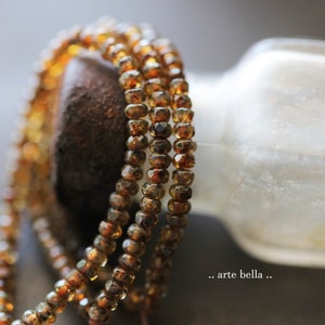 May include: Close-up of several strands of amber-colored gemstone beads. The beads are small, faceted, and strung together, creating a layered effect. The background is blurred, with a glimpse of a white bottle and the text ".. arte bella ..".