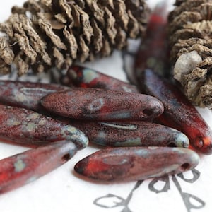 Könnte beinhalten: Nahaufnahme von mehreren länglichen, rötlich-braunen Glasperlen mit gesprenkeltem, marmoriertem Aussehen. Die Perlen haben ein Loch durch sie gebohrt. Zwei Tannenzapfen befinden sich im Hintergrund. Die Perlen sind wahrscheinlich für die Schmuckherstellung.