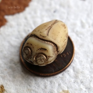 May include: A small, white, oval-shaped bead with gold-colored details. The bead is sitting on top of a penny.