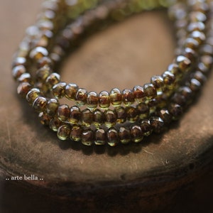 May include: Close-up of three strands of olive green and brown beads. The beads are small, faceted, and strung together. The image is slightly out of focus, with the beads in the foreground being the sharpest. The text ".. arte bella .." is in the bottom left corner.