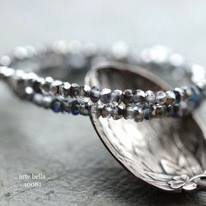 May include: Two strands of silver and blue glass beads on a silver spoon. The beads are faceted and have a shimmery finish. The spoon has a decorative design.