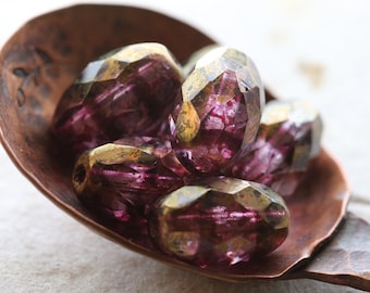 May include: A group of purple glass beads with gold accents, displayed in a copper spoon. The beads are faceted and have a translucent appearance.