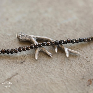 May include: A necklace with small, dark brown beads is draped over a silver-colored antler-shaped pendant. The pendant has a textured surface and is resting on a textured, neutral-toned surface. The text ".. arte bella .." and "11326" is visible.