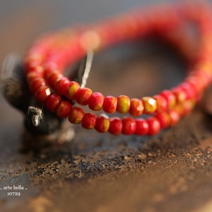 May include: A close-up of a red and yellow beaded necklace. The beads are small and round, and they are strung together in a simple pattern. The necklace is lying on a brown surface.