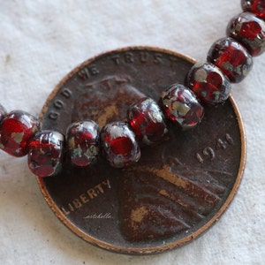 May include: A strand of red glass beads with silver flecks. The beads are round and have a slightly irregular shape. The beads are lying on top of a US penny with the year 1941 visible.