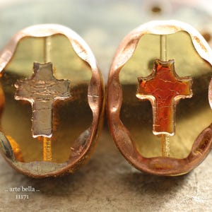 May include: Two oval-shaped beads with a gold-colored background and a copper-colored rim. Each bead features a cross design in the center. The text ".. arte bella .. 11171" is visible at the bottom of the image.