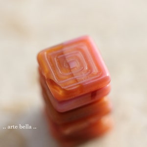Può includere: Pila di quattro piastrelle di vetro quadrate con un motivo a spirale in tonalità rosa e arancio.