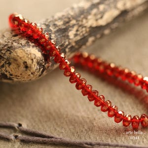 May include: A string of red faceted beads, possibly gemstones, is draped over a piece of weathered wood. The beads are strung together, creating a delicate, shimmering effect. The image includes the text "arte bella" and the number "11341".