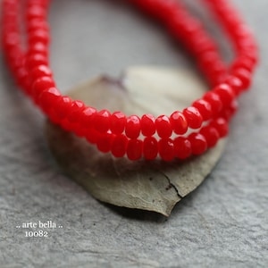 May include: A close-up of a strand of bright red glass beads. The beads are faceted and have a shiny finish. The beads are arranged in a double strand and are lying on a light brown surface.